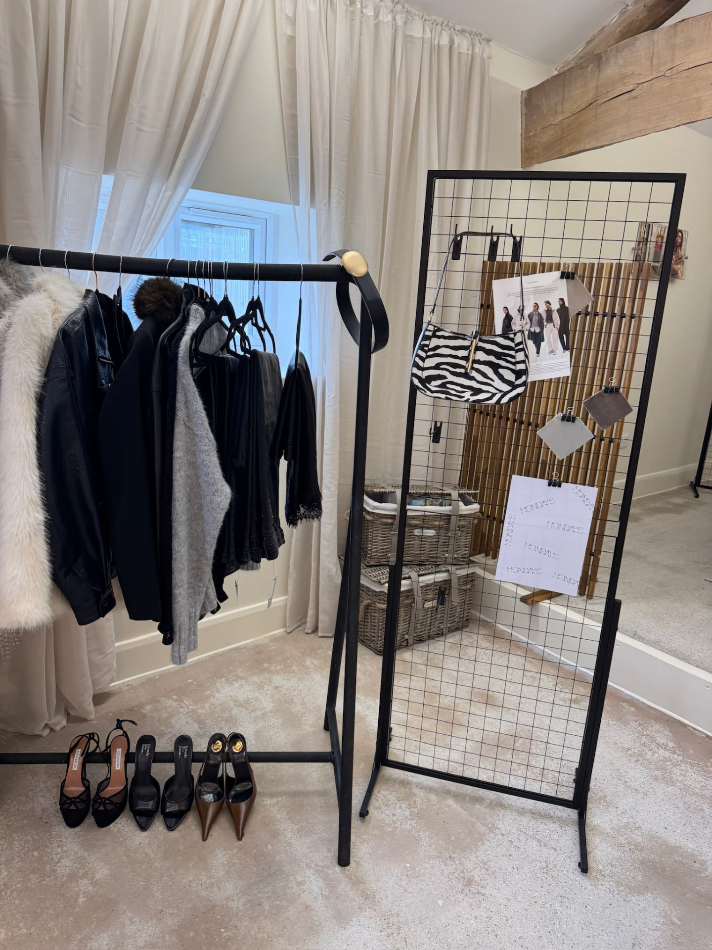 A clothing rack with a faux-fur coat, leather jacket and heels against the sheer drapes at Mondays Content Co